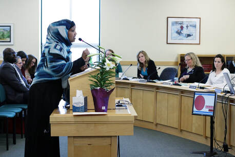 A photograph of a Black woman wearing a hijab standing in front of a panel of legislators, speaking into a microphone.